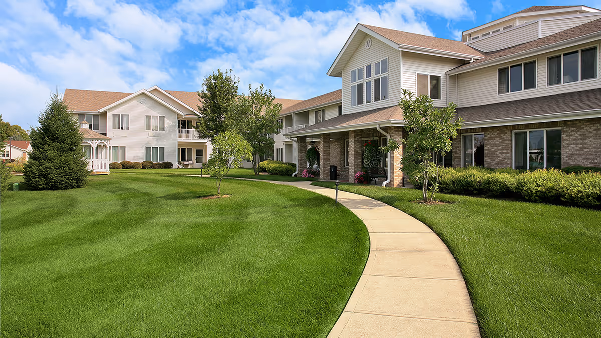 Curved sidewalk leads through a manicured lawn to the front entrance of a two-story senior living building under a blue sky.