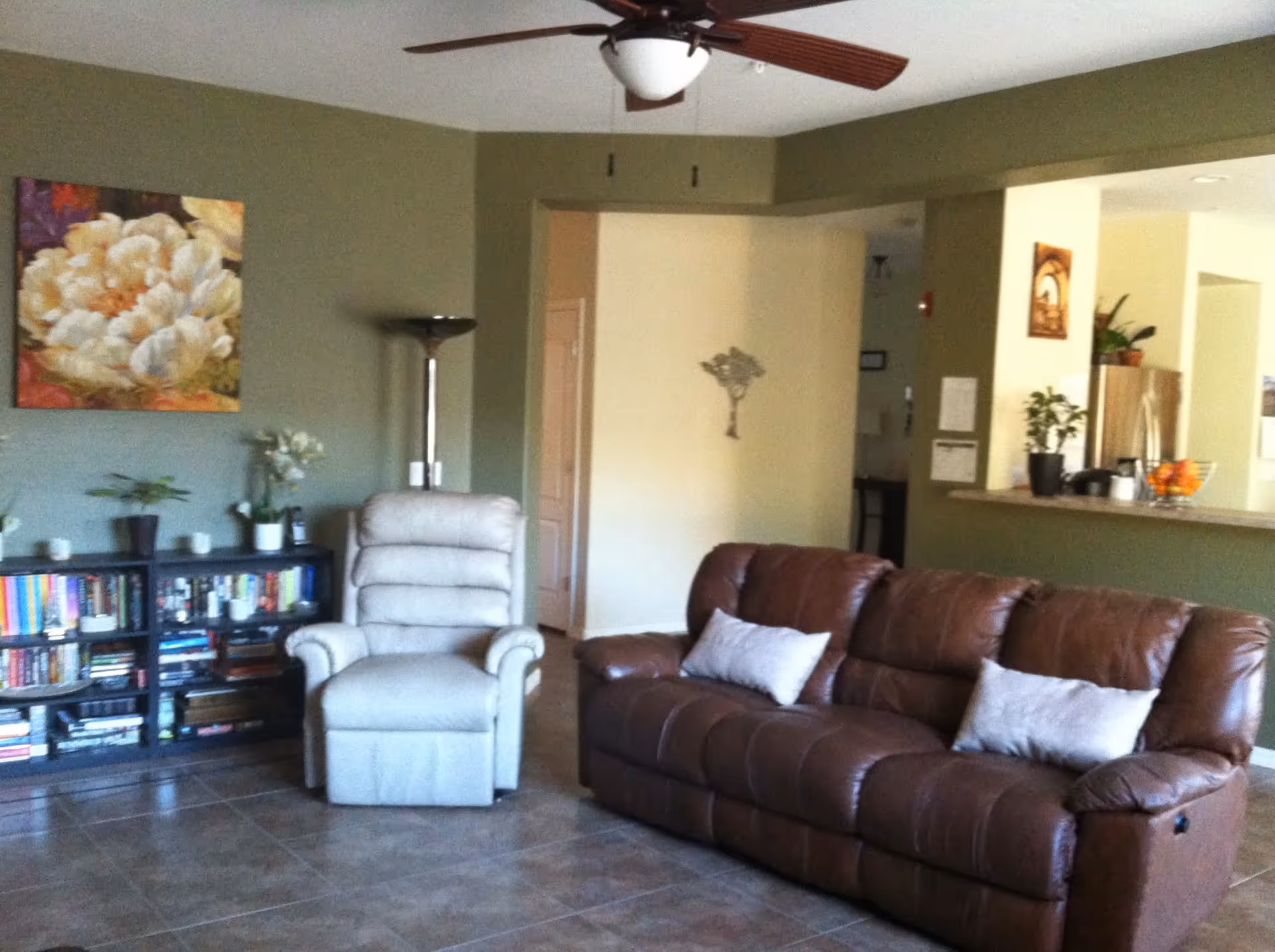 A living room with a brown leather sofa with two light-colored pillows, a beige recliner chair, a black bookshelf filled with books, a tall floor lamp, and a large floral painting on the green wall. The room has tiled flooring and a ceiling fan with wooden blades. An open kitchen area with a counter and some plants is visible in the background.