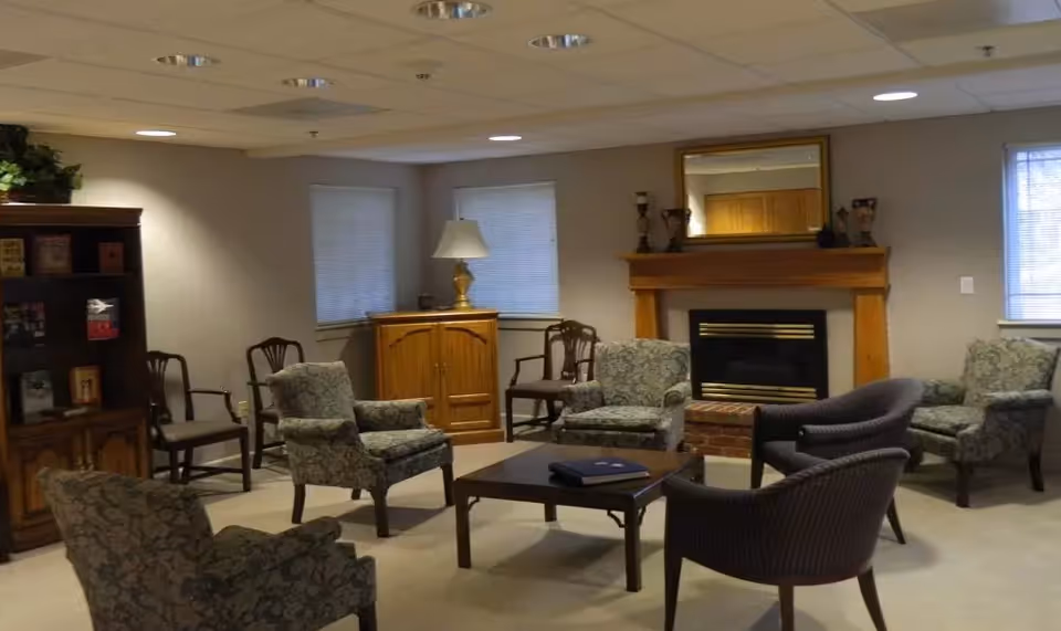 Cozy common lounge with upholstered chairs arranged around a coffee table facing a fireplace and bookcase.