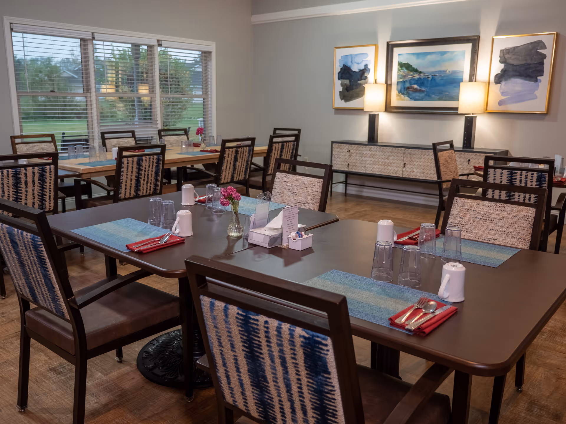 A dining room with multiple tables set with placemats, glasses, napkins, and utensils. The room has large windows with blinds, wooden chairs with patterned upholstery, and a sideboard with two lamps and framed artwork above it.