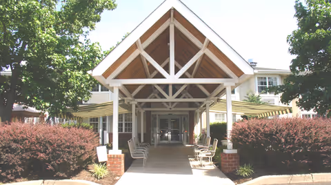 Entrance of a senior living facility with a covered walkway supported by white beams and brick pillars. There are chairs arranged along the walkway and bushes on either side. The building is light-colored with multiple windows and surrounded by trees.