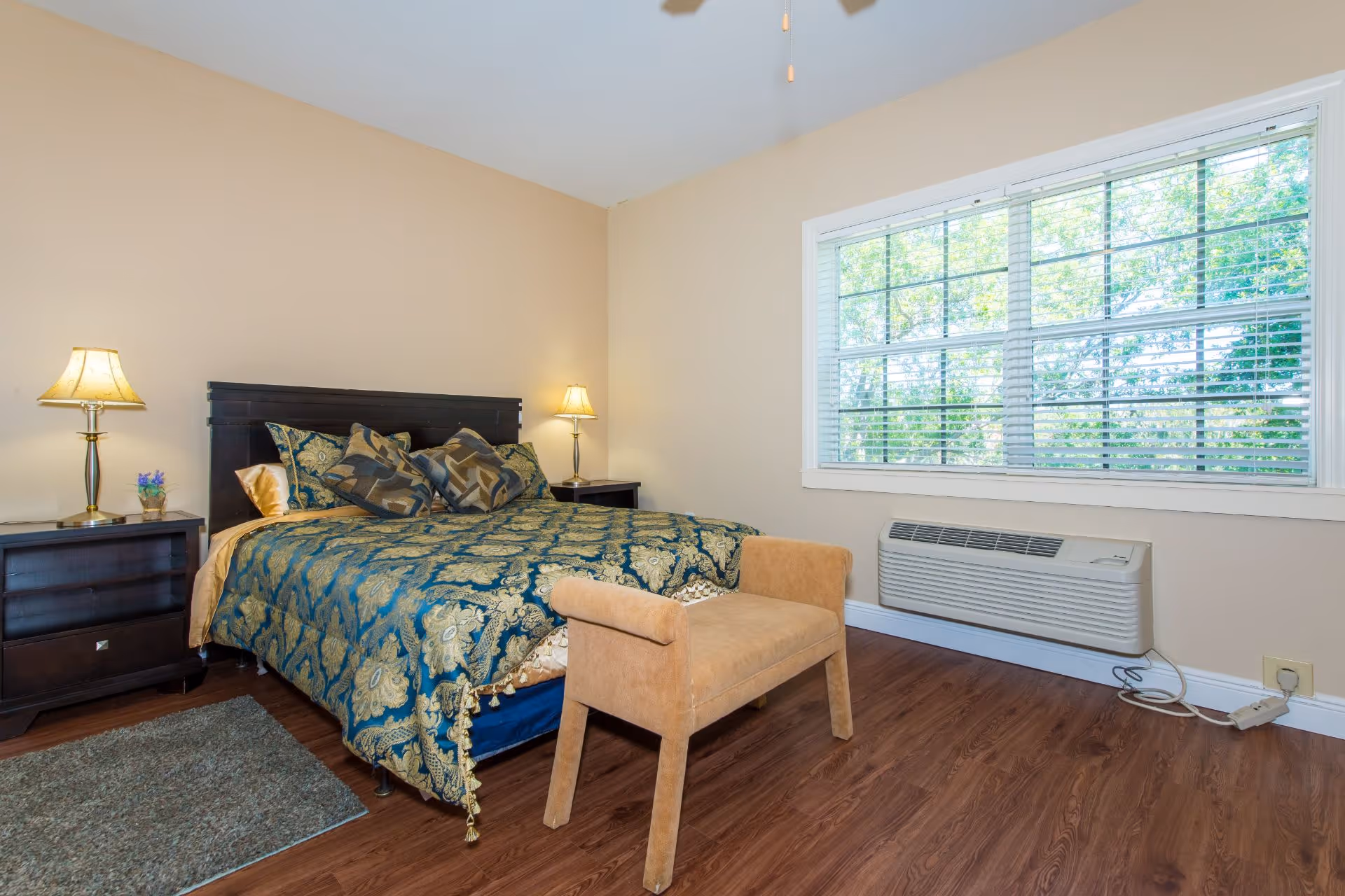 A bedroom with a large bed covered in a blue and gold patterned comforter and multiple pillows. There are two dark wooden nightstands on either side of the bed, each with a lamp. A beige upholstered bench is placed at the foot of the bed. The room has wooden flooring, a large window with white blinds, and a wall-mounted air conditioning unit below the window.