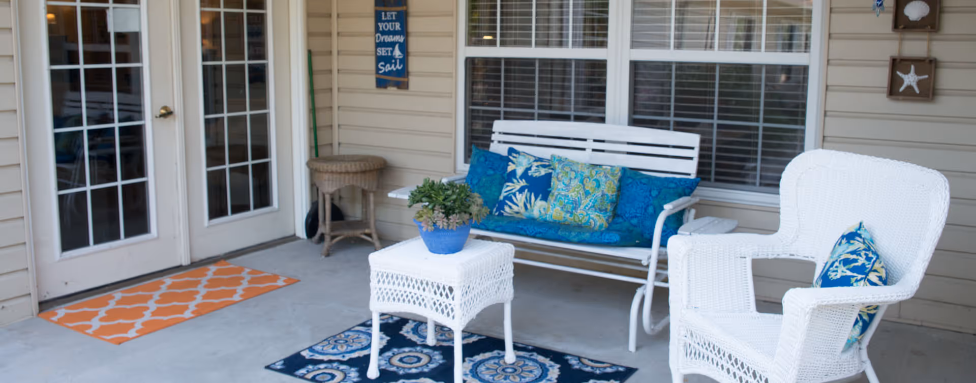 Outdoor patio area with white wicker furniture including a bench with blue and patterned cushions, a chair with a blue cushion, and a small table with a potted plant. The patio has a concrete floor with two decorative rugs, beige siding walls, a double glass door, and a window with white blinds. Nautical-themed wall decorations are visible.