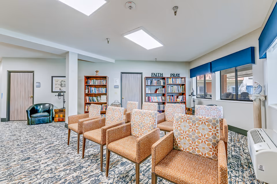 Well-lit senior living common room with patterned armchairs arranged in rows, bookshelves labeled "FAITH PRAY," and large windows.