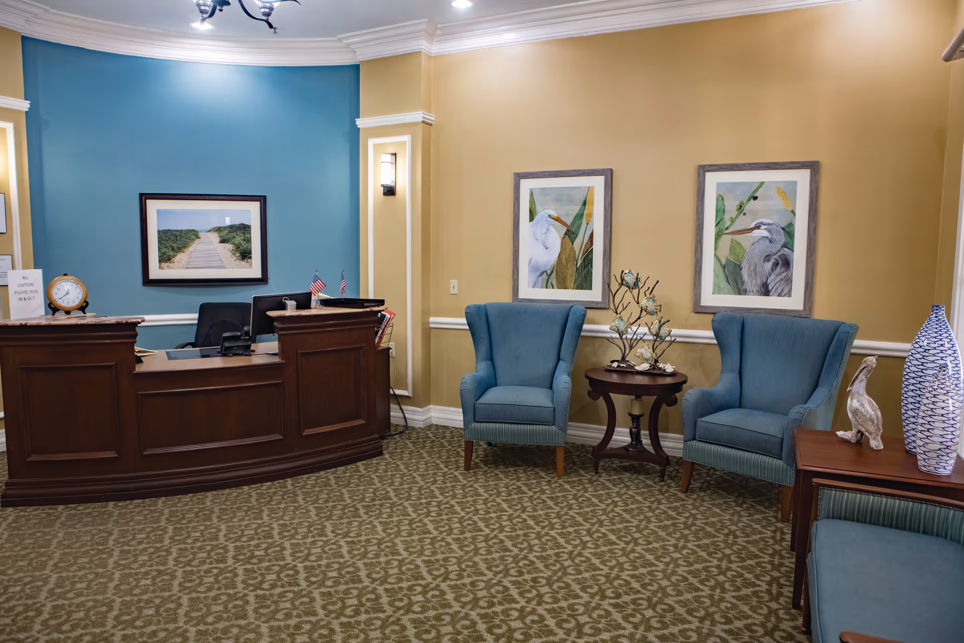 Reception area with a dark wooden desk, two blue upholstered armchairs, a small round table with decorative items, framed bird artwork on the walls, and a patterned carpet. The walls are painted in blue and beige tones.