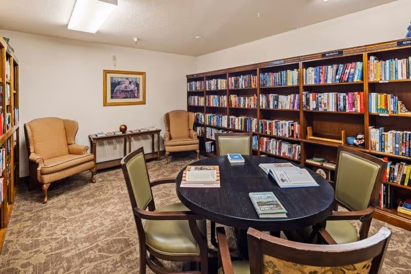 A cozy library room with bookshelves filled with books along two walls. There is a round black table in the center with four chairs around it, and several books and magazines are placed on the table. Two upholstered armchairs are positioned against the far wall beneath a framed picture. The room has carpeted flooring and soft lighting from a ceiling fixture.