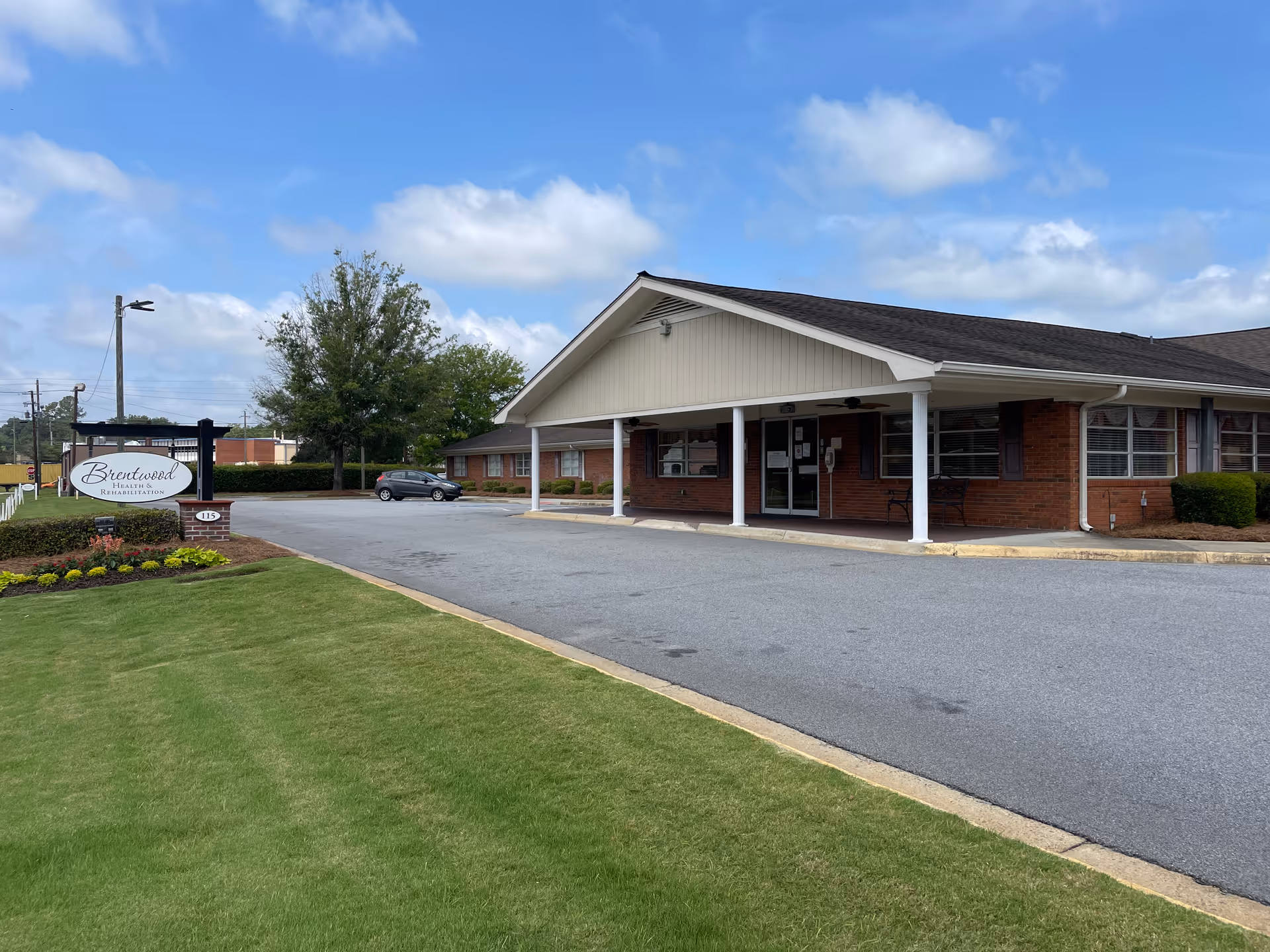 Exterior view of Brentwood Health & Rehabilitation building with a covered entrance, parking area, green lawn, and a sign displaying the facility's name under a partly cloudy sky.