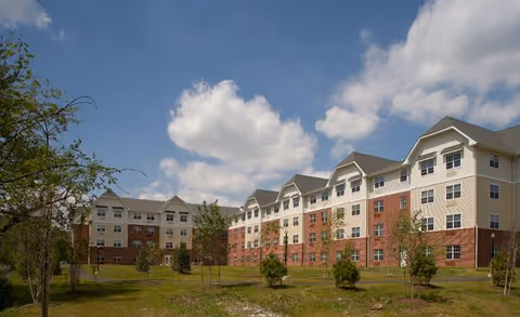 Exterior view of a multi-story residential building with a combination of brick and light-colored siding under a partly cloudy sky, surrounded by grass and small trees.