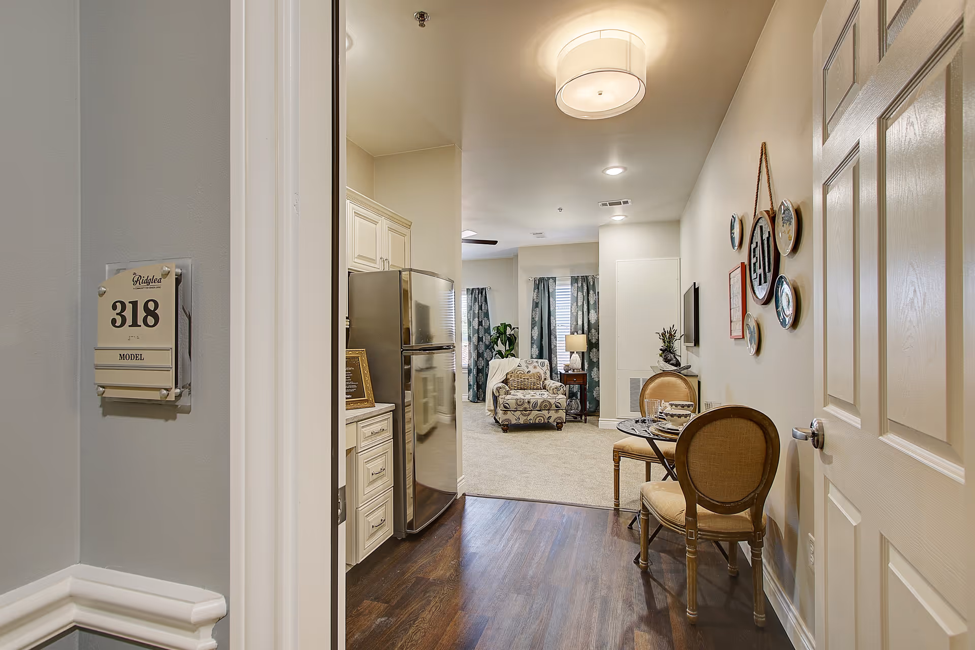 View into a senior living apartment unit labeled 318 at The Ridglea Senior Living, showing a small dining area with two chairs and a table set for two, a kitchen area with a stainless steel refrigerator, and a living room with an armchair, side table, lamp, and window with curtains.