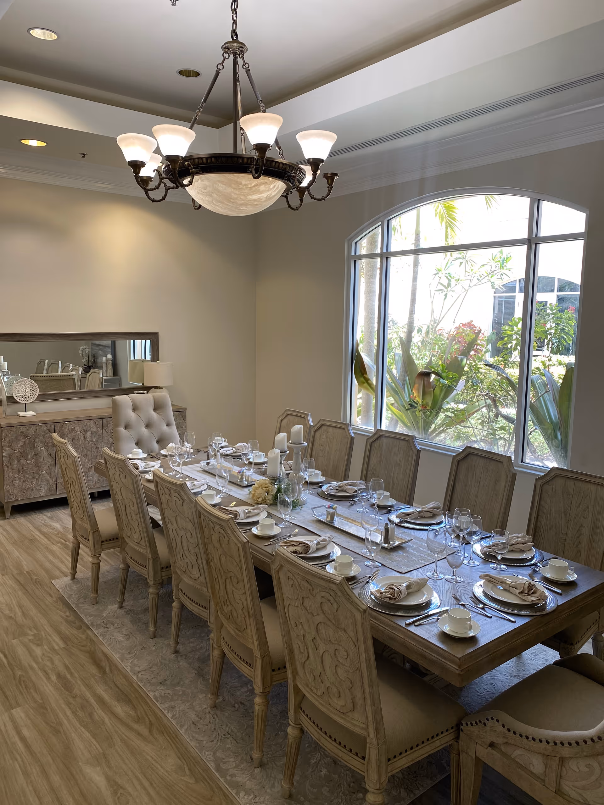 A formal dining room with a long wooden table set for twelve people. The table is arranged with plates, napkins, cups, wine glasses, and silverware. There is a decorative centerpiece with candles and flowers. The room has a large window with a view of greenery outside, a chandelier hanging from the ceiling, and a sideboard with a lamp and decorative items.