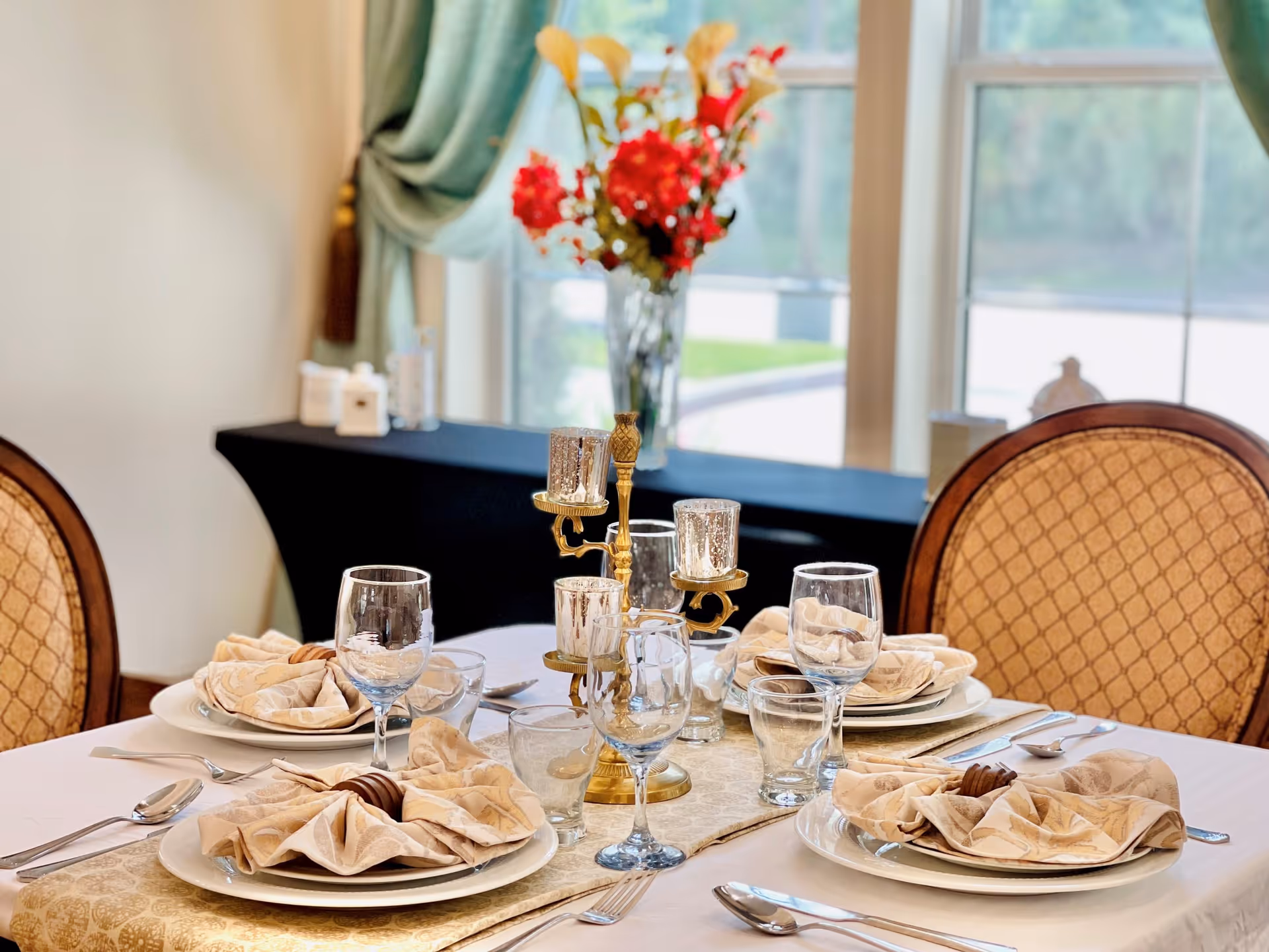 A dining table set for four with beige patterned napkins, plates, glasses, and silverware. A gold-colored candle holder with four candles is in the center of the table. In the background, there is a window with green curtains and a vase with red and white flowers on a black table.