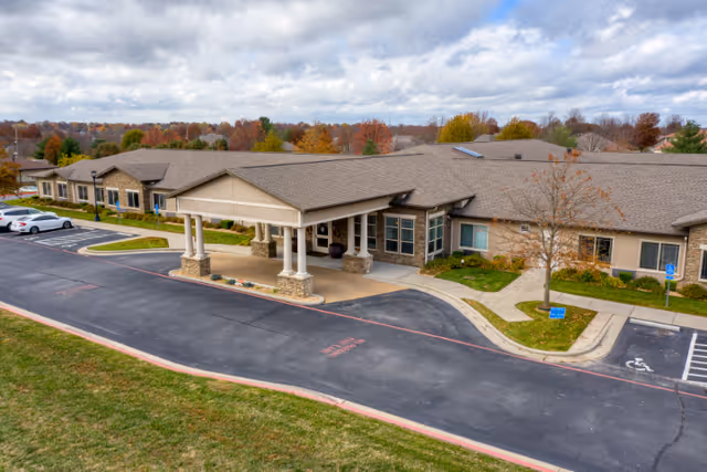 Exterior view of Cedarhurst Senior Living of Springfield, MO, showing a single-story building with a covered entrance supported by columns. The surrounding area includes a parking lot with several cars, a sidewalk, and some trees with autumn foliage under a cloudy sky.