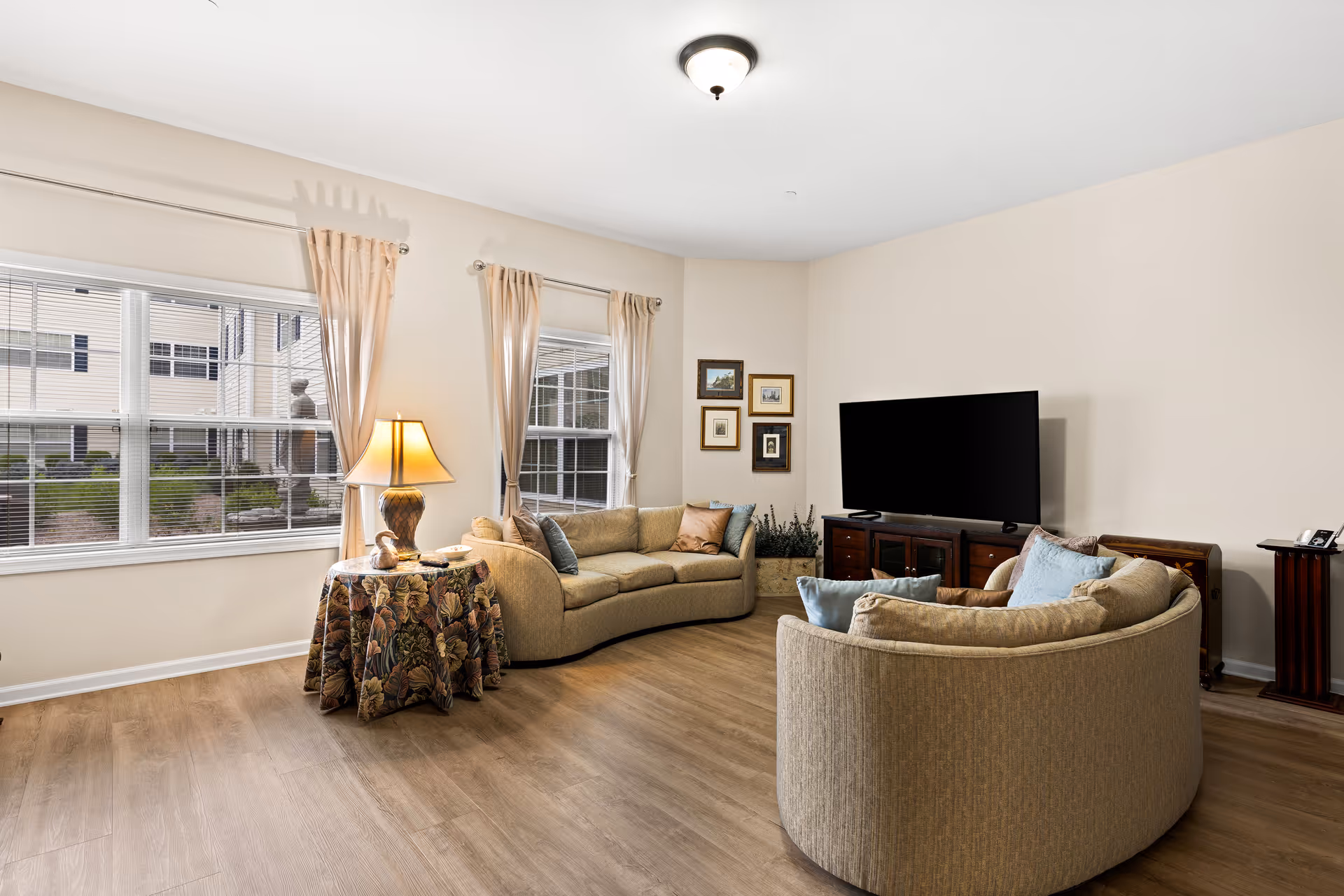 A bright living room with two curved beige sofas adorned with blue and beige pillows, a wooden TV stand with a flat-screen television, a side table covered with a floral tablecloth and a lamp, and three windows with beige curtains letting in natural light.