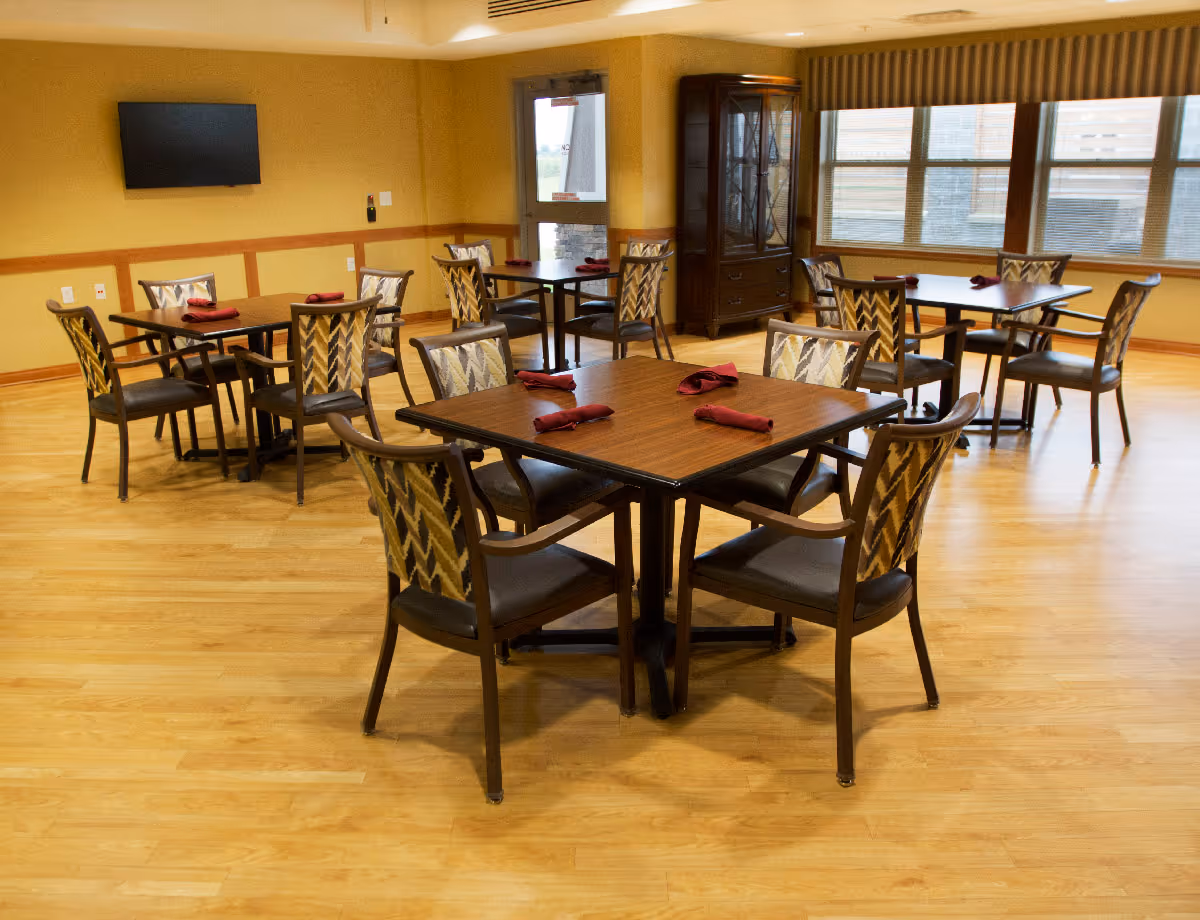 A dining room with several square wooden tables, each surrounded by four chairs with patterned backs and dark seats. Each table has neatly folded red napkins placed on it. The room has light wooden flooring, beige walls with wooden trim, a wall-mounted flat screen TV, a glass-front cabinet, and large windows with blinds letting in natural light.