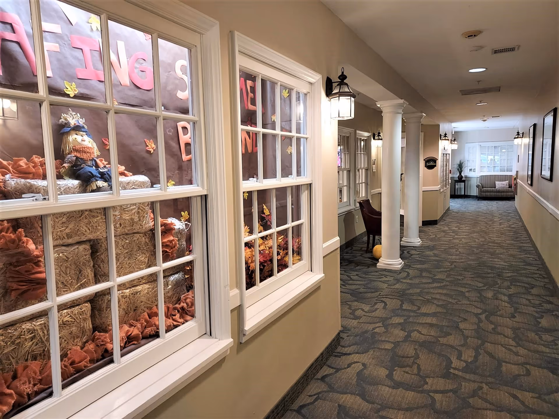 A well-lit hallway in a senior living facility with beige walls, patterned carpet, white columns, and wall-mounted lantern-style lights. On the left side, there are windows displaying a fall-themed decoration with hay bales, a scarecrow doll, and autumn leaves. At the end of the hallway, there is a seating area with a striped couch and a small table with a plant.