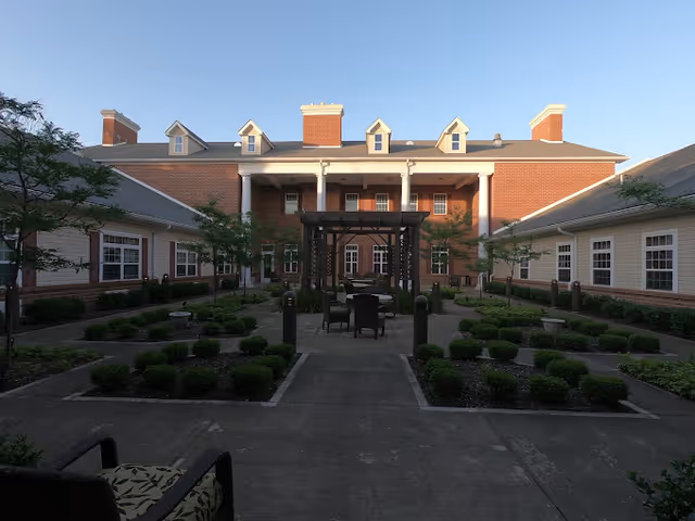 Outdoor courtyard area of a senior living facility with neatly trimmed bushes, paved walkways, and a pergola with chairs and tables in the center. The courtyard is surrounded by a two-story brick building with white columns and multiple windows.