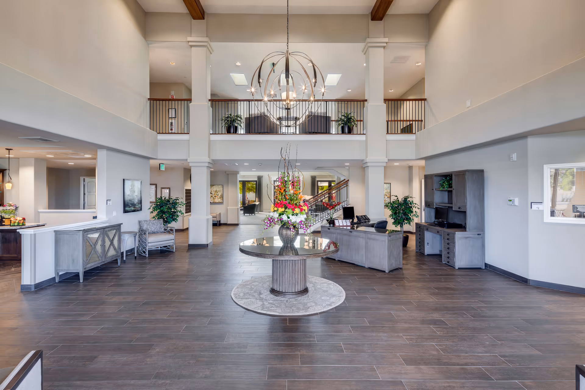 Spacious and well-lit senior living facility lobby with high ceilings, a large round table with a floral arrangement in the center, seating areas, a reception desk, and a staircase leading to an upper balcony with additional seating and plants.