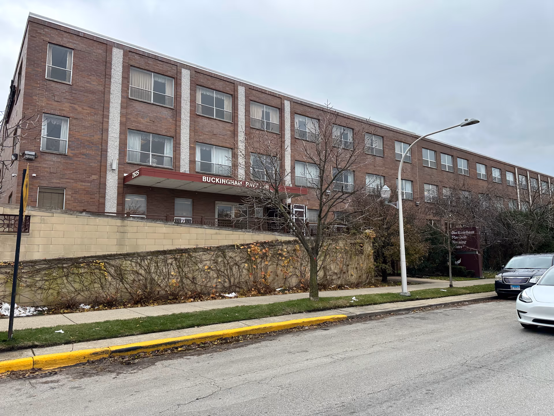 Exterior view of Buckingham Pavilion Nursing and Rehabilitation, a three-story brick building with multiple windows. There is a sidewalk and street in front with a few parked cars, leafless trees, and a cloudy sky overhead.