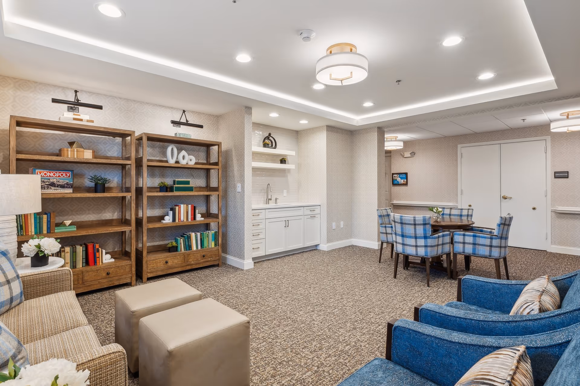 Brightly lit senior living common room with bookshelves, seating, a small kitchenette and a dining table with plaid chairs.