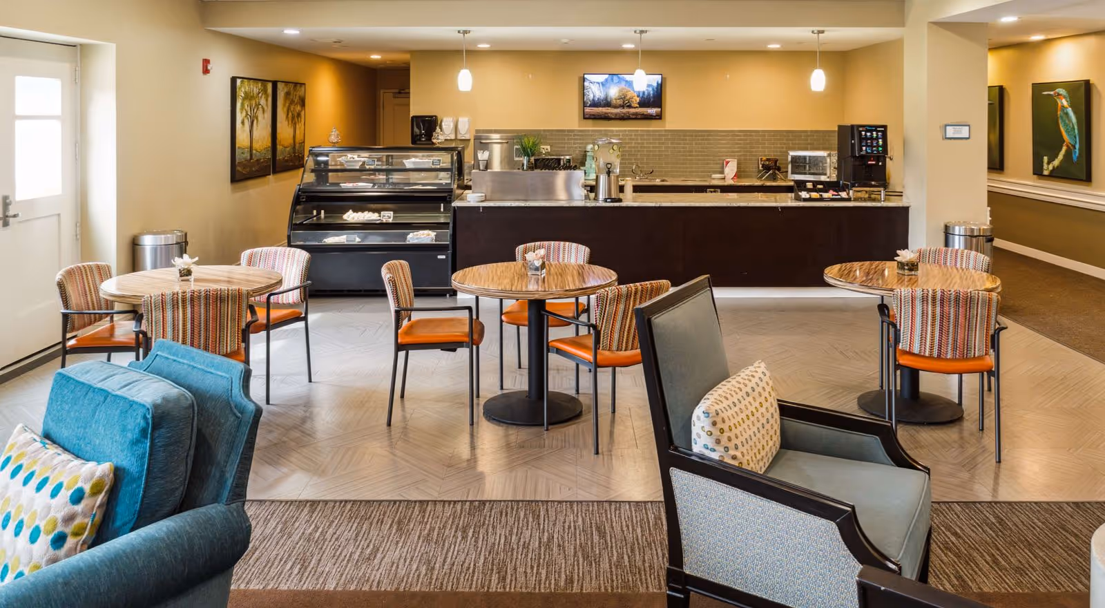 Bright communal dining area with round tables and upholstered chairs, lounge seating in the foreground, and a service counter with display cases in the back.