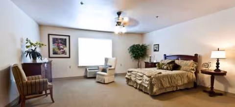 Spacious senior living bedroom with a bed, armchairs, dresser, lamps, and large window.