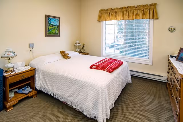 A cozy bedroom with a double bed covered in a white textured bedspread and a red blanket folded at the foot. There is a small teddy bear on the bed. The room has beige walls, a window with blinds and a yellow valance, a wooden nightstand with a vintage lamp and tissues, a dresser with framed photos, and a framed painting on the wall above the bed.