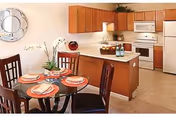 A kitchen and dining area in an apartment with wooden cabinets, white appliances including a refrigerator, stove, and microwave. A round glass dining table is set with four chairs, each place setting having a plate, napkin, and utensils. A decorative mirror hangs on the wall and a plant is placed on the dining table.