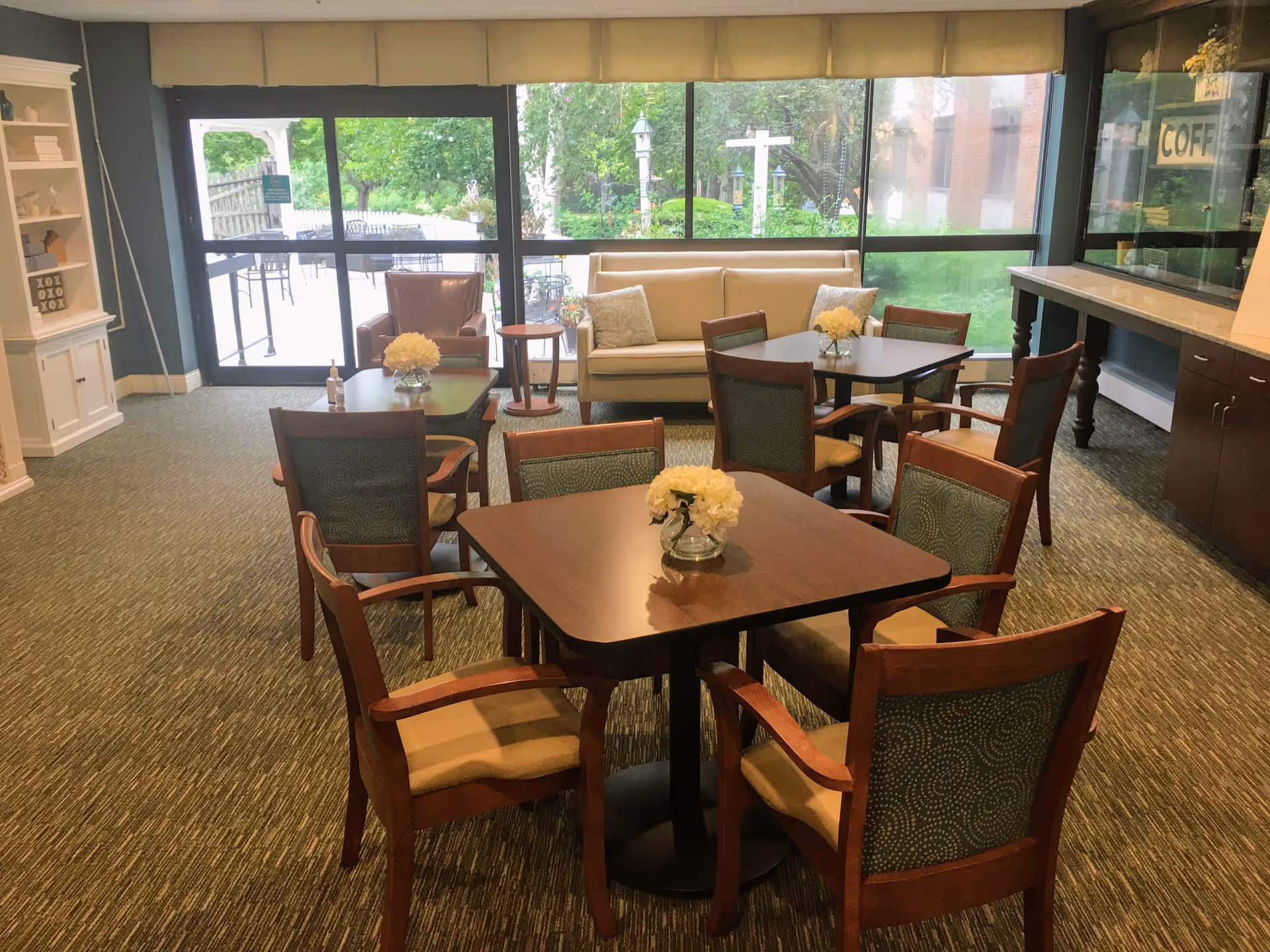 Communal dining/lounge area with several small tables and chairs, a sofa and armchair, and large windows overlooking a garden.