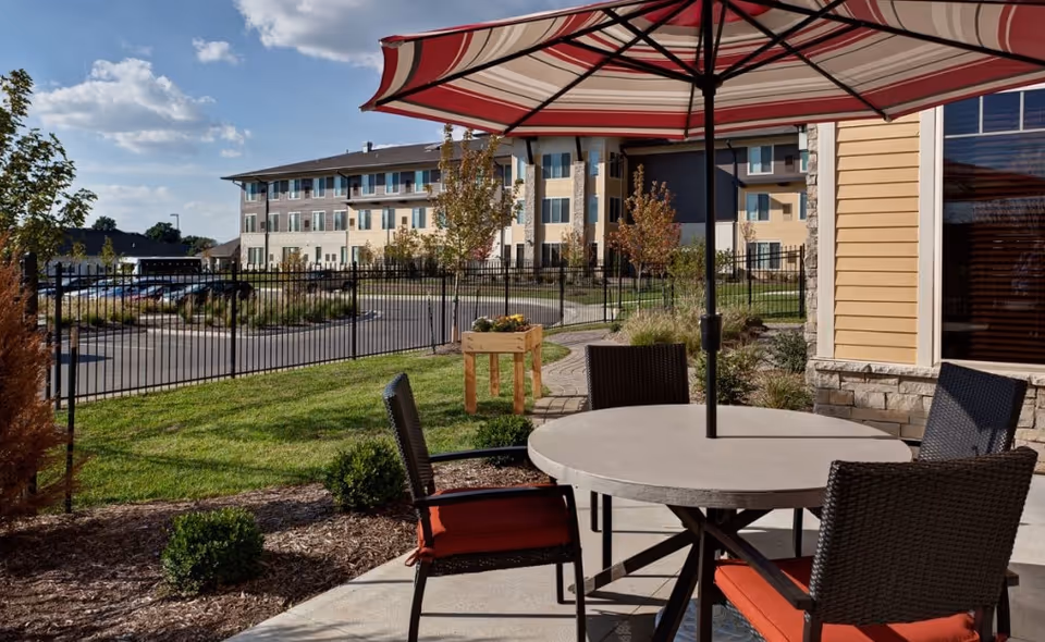 Outdoor patio area with a round table and four wicker chairs with red cushions under a large striped umbrella. In the background, there is a fenced grassy area with small bushes and trees, and a multi-story building under a partly cloudy sky.