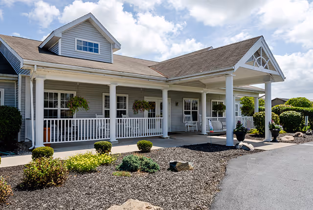 Exterior view of a single-story building with a covered porch supported by white columns. The porch has white railings and hanging flower baskets. There are shrubs and landscaping with mulch in front of the building under a partly cloudy sky.