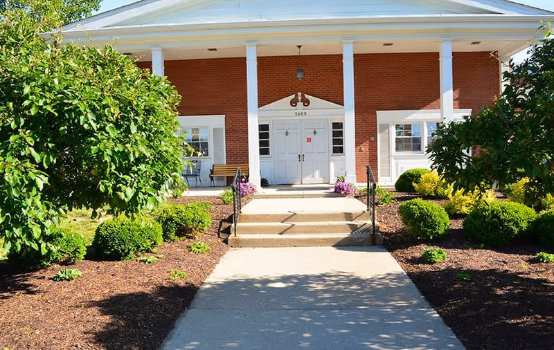 Front entrance of a brick building with white columns and double white doors, surrounded by neatly trimmed bushes and trees with a concrete walkway leading up to the steps.