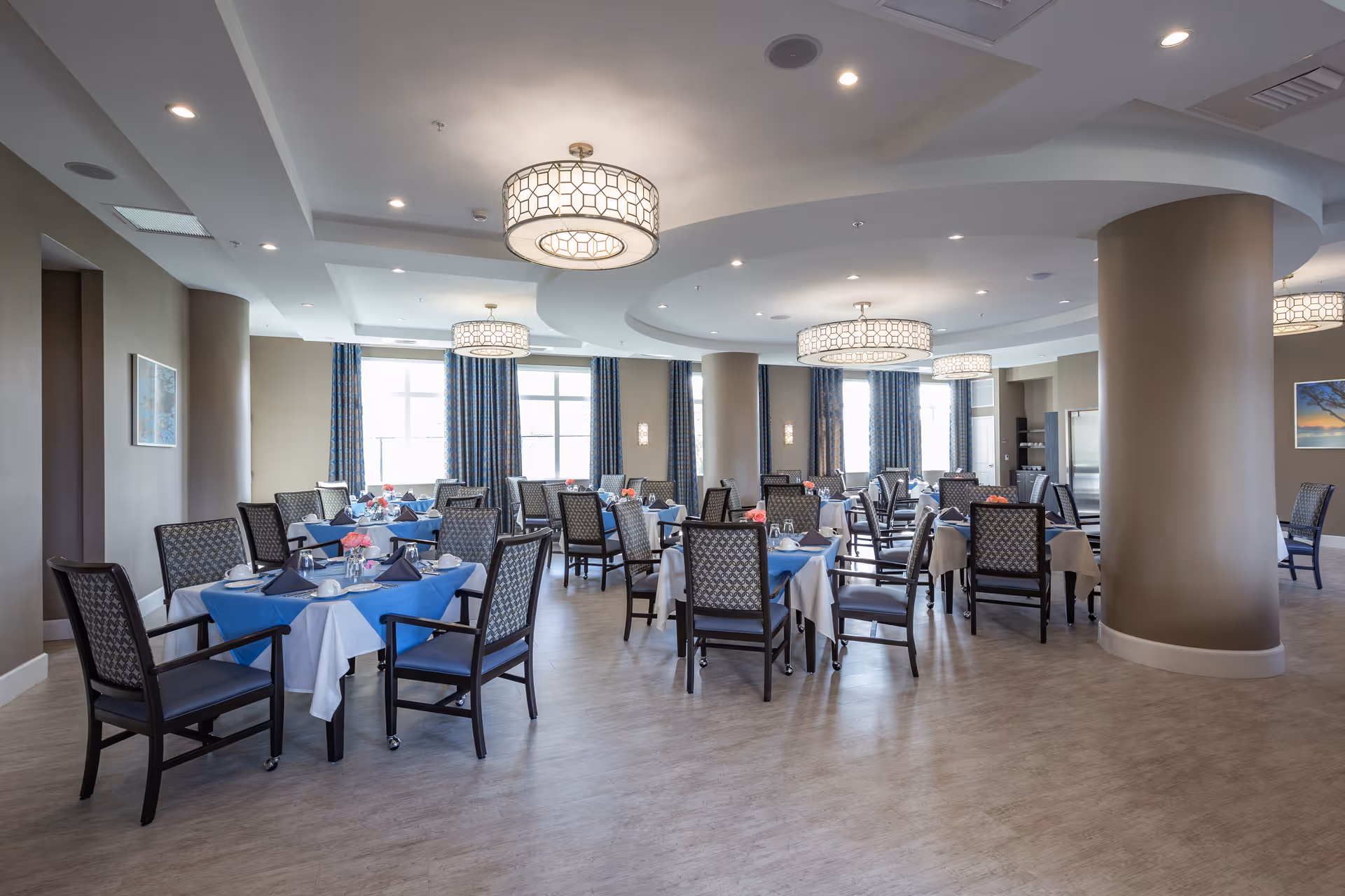 A spacious dining room with multiple tables covered in blue and white tablecloths, each set with plates, cups, and napkins. The room features large windows with blue curtains, patterned chairs, beige walls, and modern circular ceiling lights.