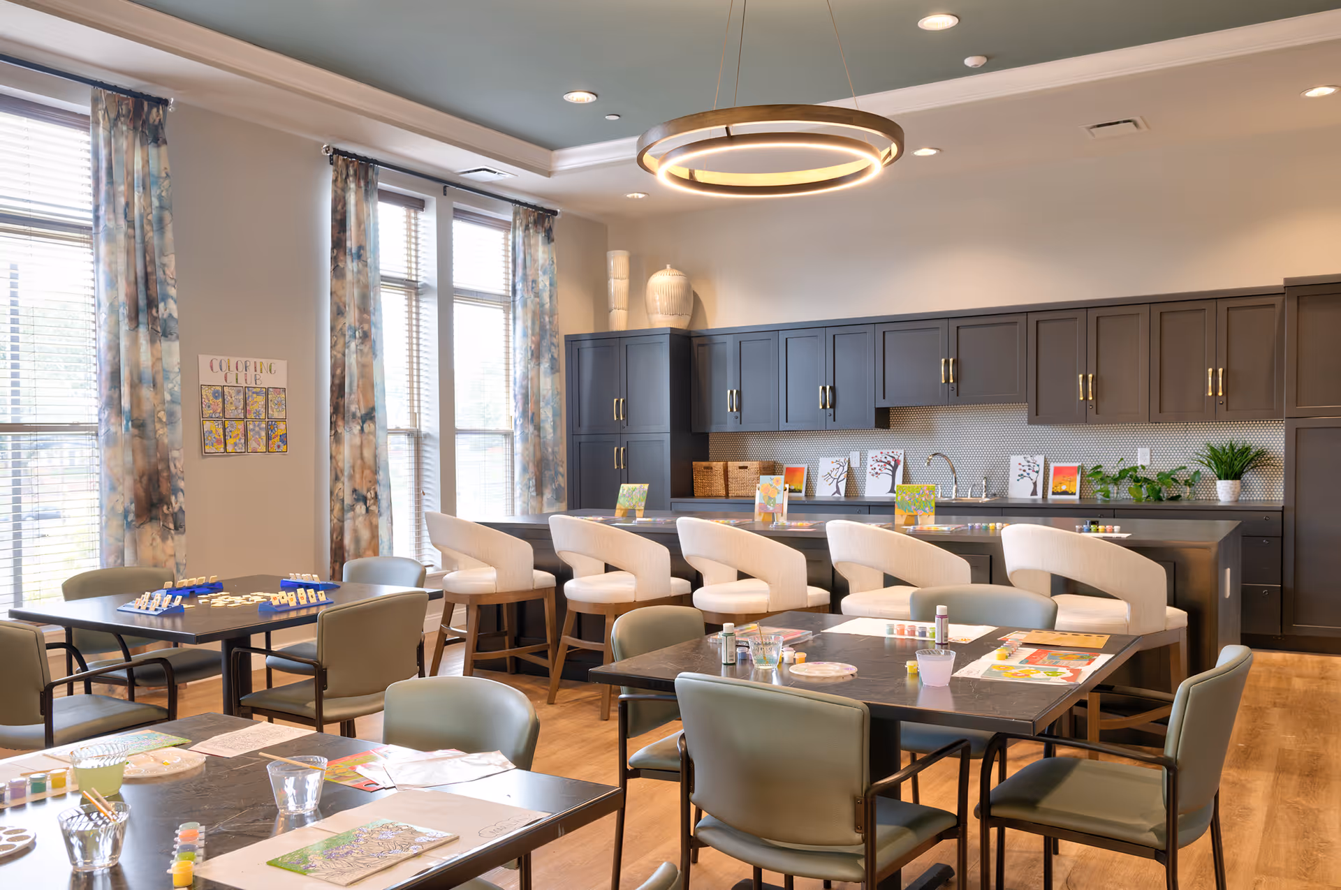 A bright and spacious activity room with large windows covered by patterned curtains. The room features several tables and chairs arranged for group activities, with art supplies and coloring materials on the tables. In the background, there is a kitchen area with dark cabinets, a countertop with bar stools, and decorative plants and artwork displayed on the counter.
