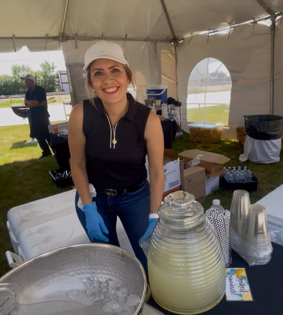 A smiling woman wearing a white cap, black sleeveless top, blue gloves, and jeans stands behind a table with a large glass dispenser filled with spiked lemonade, plastic cups, and straws under a white event tent. In the background, a man is grilling outside on the grass.