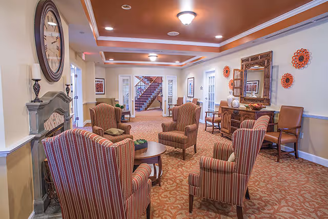 A cozy living room area in a senior living facility with four striped armchairs arranged around a round wooden coffee table. The room features warm lighting, a large wall clock above a decorative fireplace, a wooden sideboard with a mirror and decorative items, and a carpeted floor with a patterned design. In the background, there is a staircase and additional seating areas.