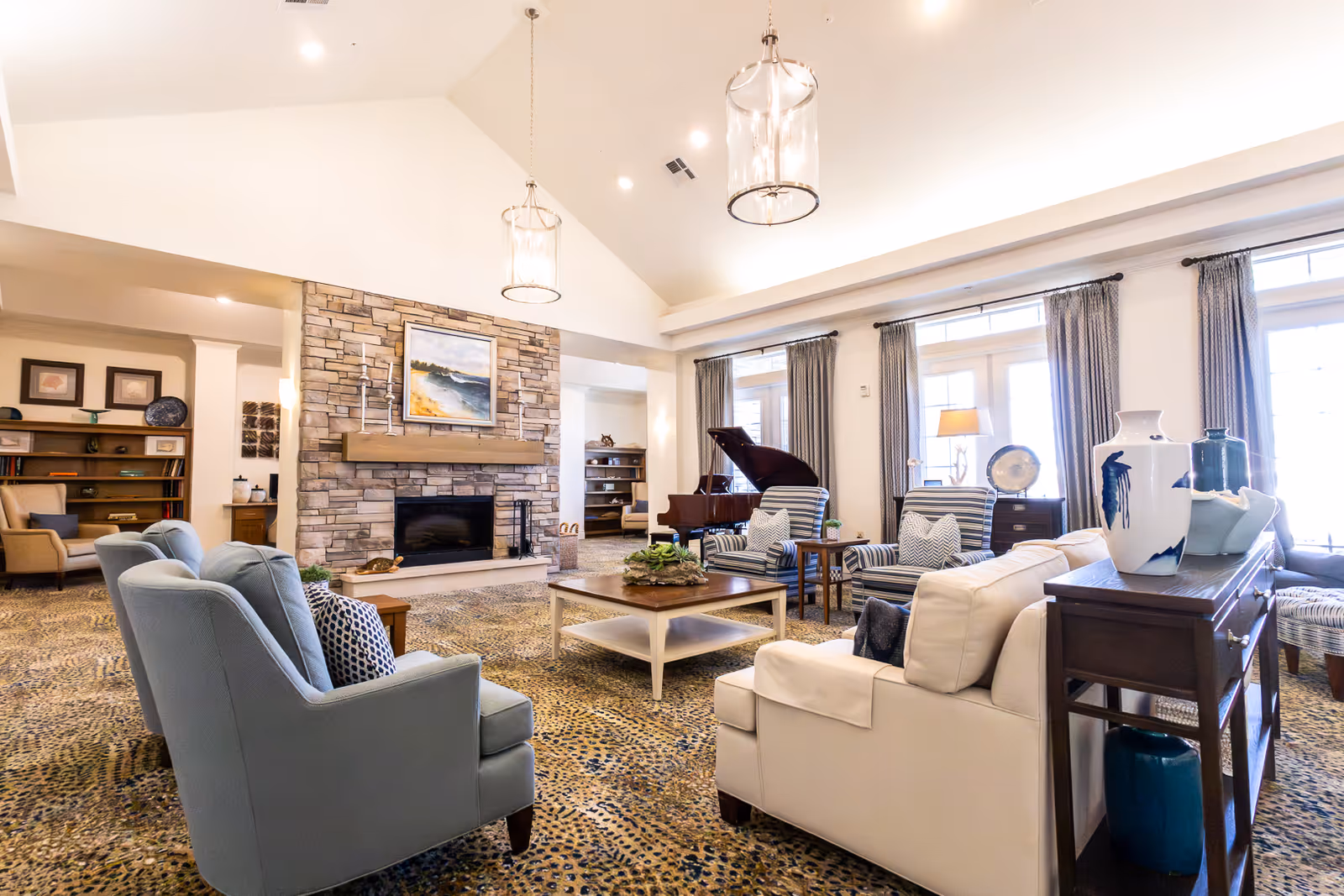 A spacious and well-lit senior living common area featuring comfortable seating including armchairs and sofas arranged around a central coffee table. The room has a high vaulted ceiling with two hanging light fixtures, a stone fireplace with a painting above it, large windows with curtains allowing natural light, a grand piano, and bookshelves with decorative items.