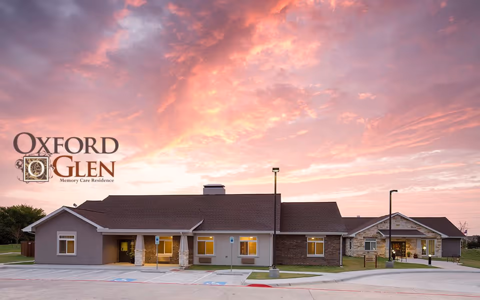 Exterior view of Oxford Glen Memory Care Residence building at sunset with a colorful sky and parking spaces in front.