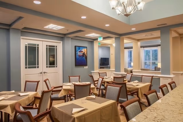 A dining room with several tables covered in beige tablecloths and set with napkins and placemats. The room has wooden chairs with cushioned seats and backs. There are double doors with glass panels on one wall, a painting hanging next to the doors, and a chandelier hanging from the ceiling. The room has soft lighting and a warm, inviting atmosphere.