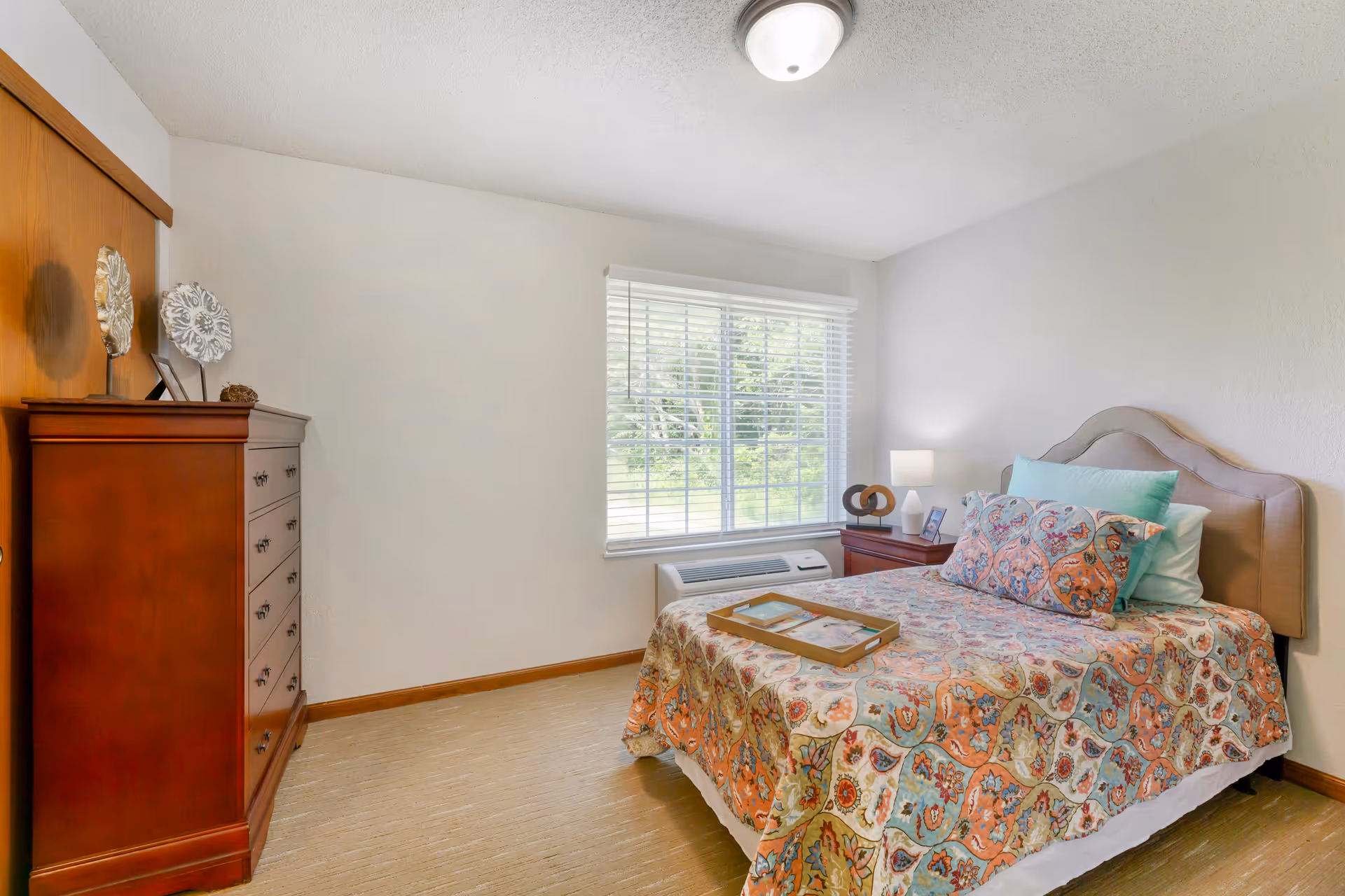 A cozy bedroom with a bed covered in a colorful patterned quilt and multiple pillows. There is a wooden nightstand with a lamp and decorative items next to the bed. A large window with white blinds lets in natural light. On the left side, there is a tall wooden dresser with decorative pieces on top. The walls are painted white and the floor has a light-colored carpet.