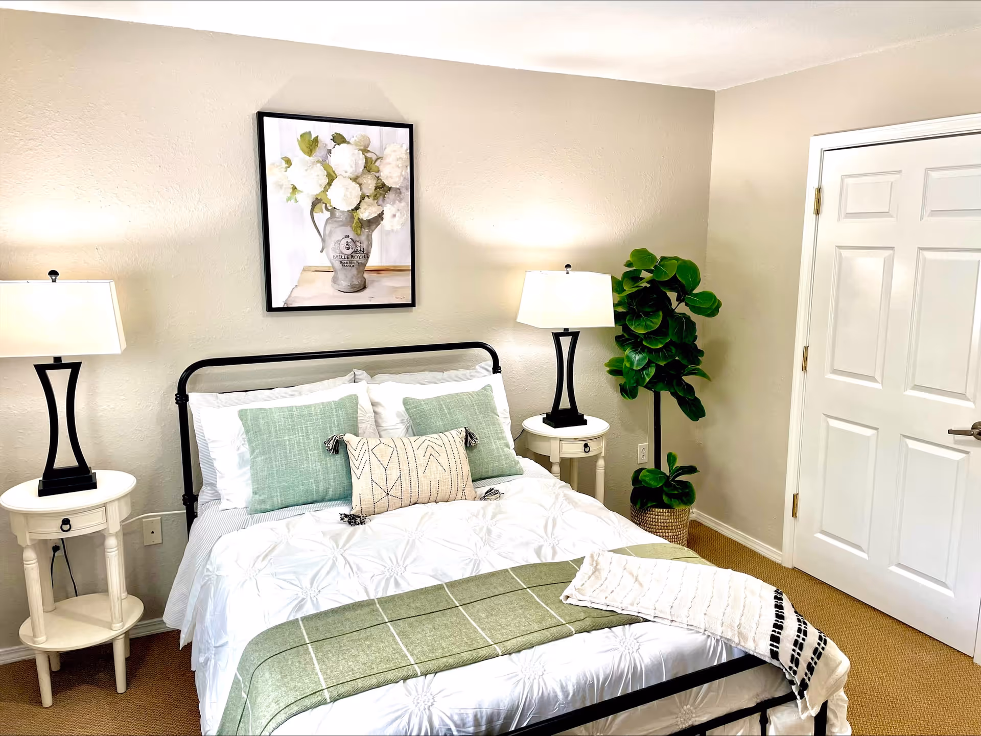 A neatly made bed with white bedding and green accent pillows in a bedroom. There are two white round bedside tables with black lamps on each side of the bed. A framed picture of white flowers in a vase hangs on the beige wall above the bed. A green potted plant is placed in the corner near a white door.
