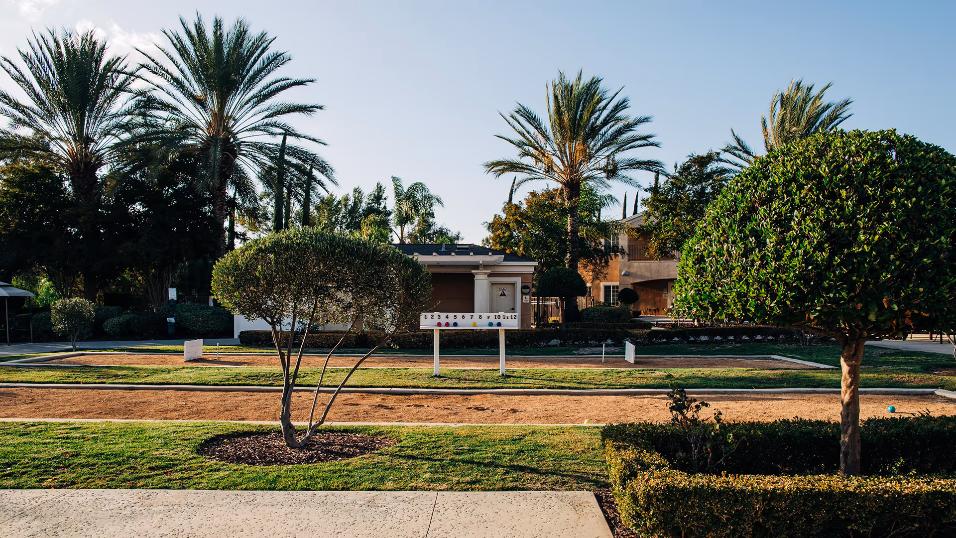Landscaped outdoor courtyard with trimmed topiary, palm trees and bocce-style courts in front of a residential building.