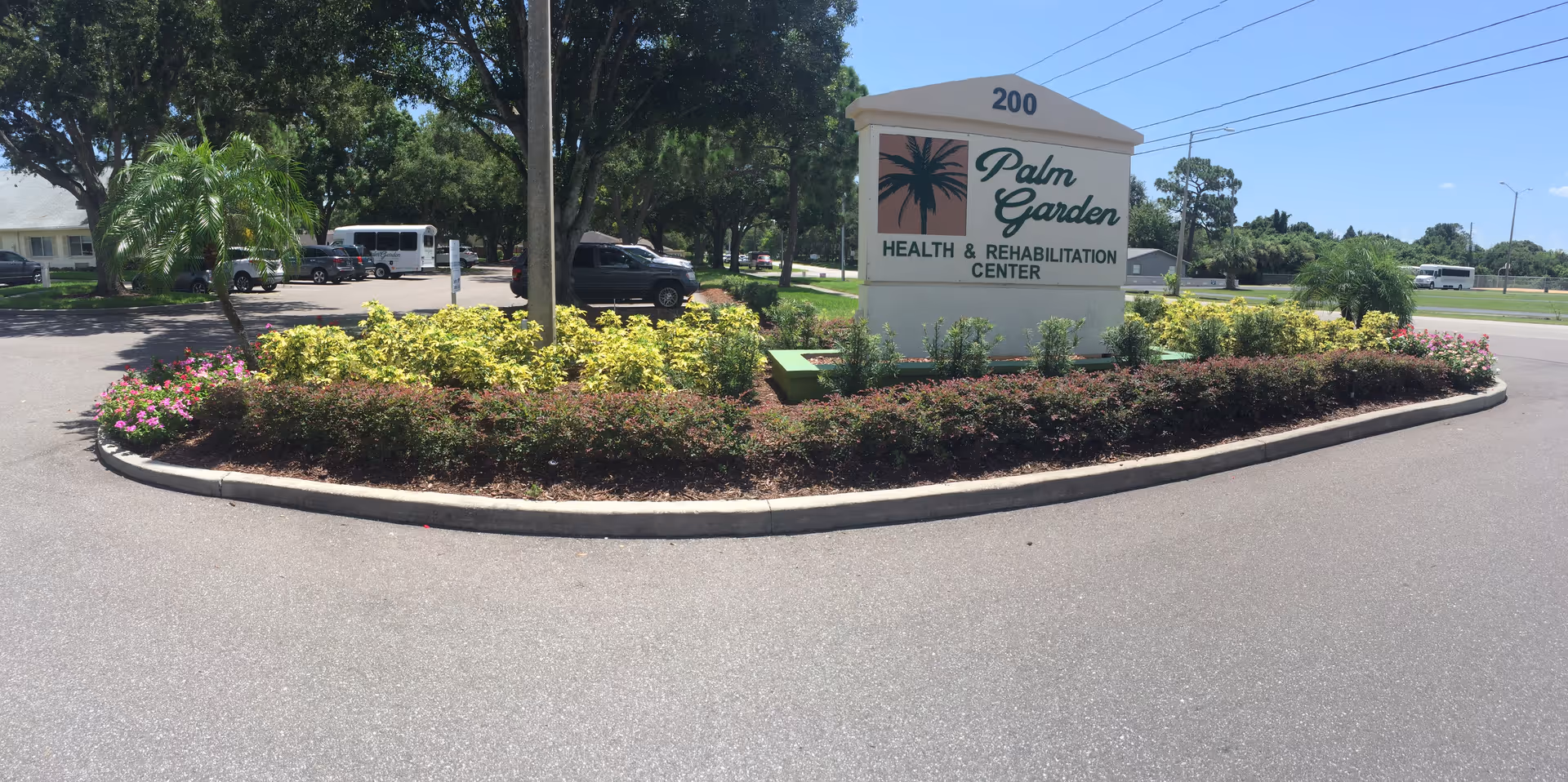 Outdoor view of the entrance area to Palm Garden Health & Rehabilitation Center featuring a large sign with the facility name and logo surrounded by landscaped bushes and flowers, with trees, parked vehicles, and a clear blue sky in the background.