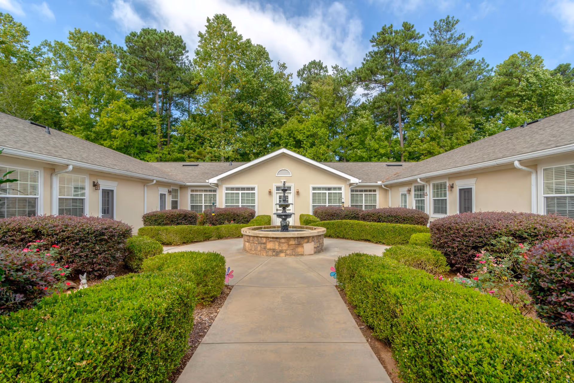 A courtyard area at Addington Place of Johns Creek featuring a central stone fountain surrounded by neatly trimmed green hedges and bushes. The courtyard is enclosed by a single-story beige building with multiple windows and doors, set against a backdrop of tall green trees under a partly cloudy sky.