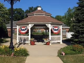 White wooden gazebo decorated with patriotic bunting and potted flowers on a paved walkway in a landscaped outdoor area.