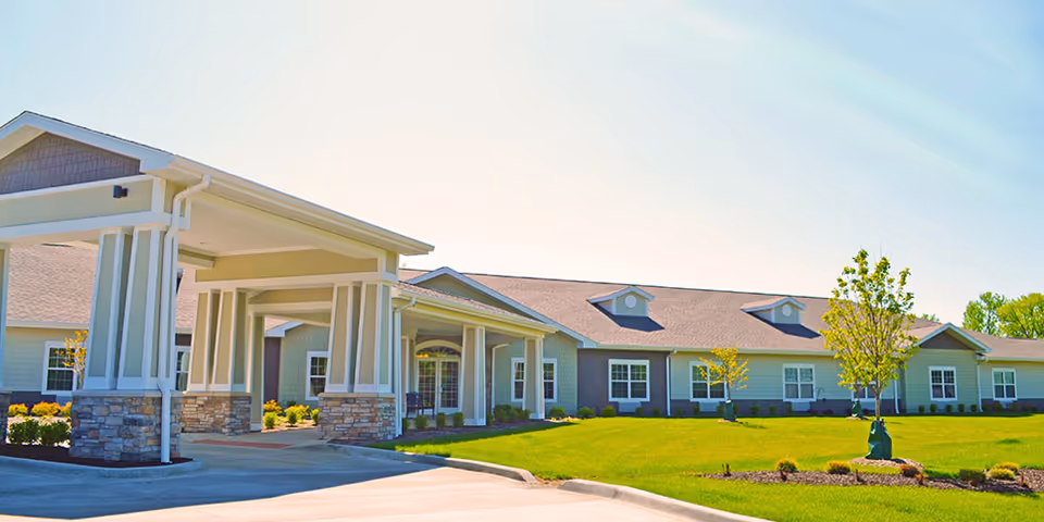 Exterior view of a single-story senior living facility building with a covered entrance supported by columns, surrounded by a well-maintained lawn and small trees under a clear sky.