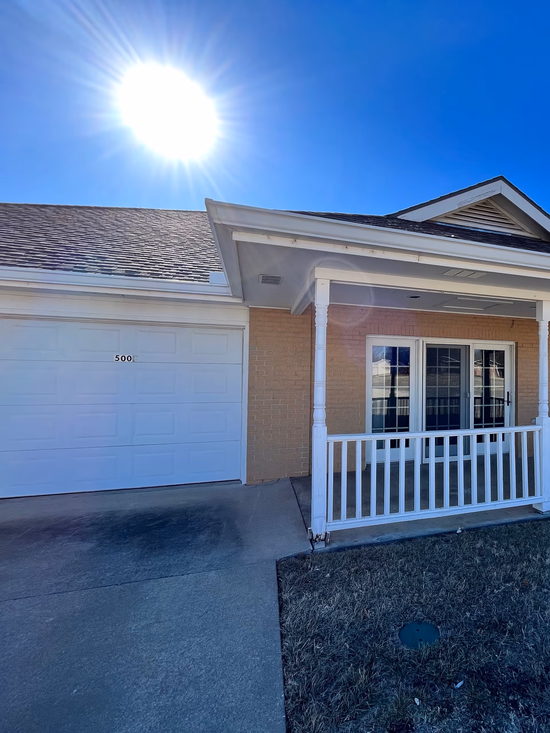 Front exterior of a single-story brick house showing a white garage door, covered porch with railing, and the bright sun in a clear blue sky.
