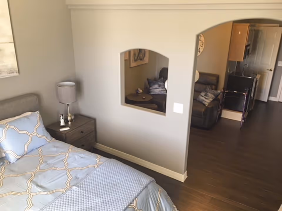 A furnished bedroom with a bed, nightstand and lamp opening into a living area and kitchenette through an arched interior wall.