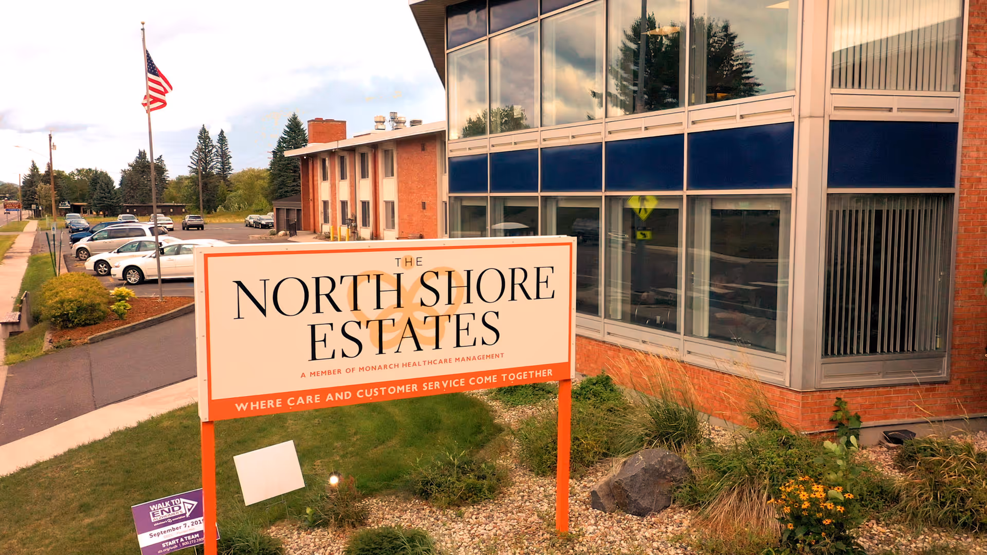 Exterior view of The North Shore Estates facility showing a large sign with the facility name and slogan, an American flag on a pole, a parking lot with several cars, and part of the building with large windows and brick walls.