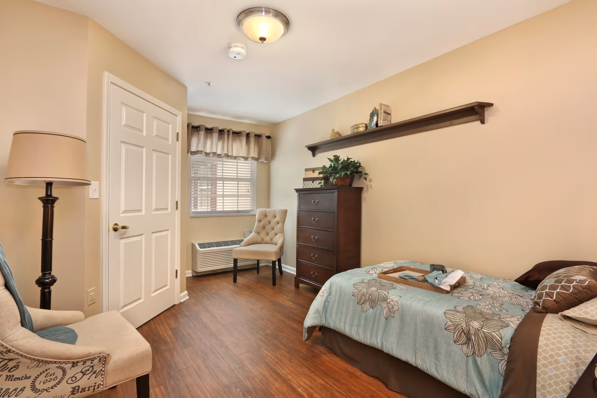 A cozy bedroom with a single bed covered in a floral patterned comforter, a wooden chest of drawers with decorative items on top, two upholstered chairs, a floor lamp, a window with beige curtains, and a wooden floor.
