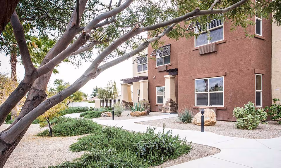 Exterior view of a two-story assisted living community building with brown and beige walls, multiple windows, and a landscaped garden featuring bushes, rocks, and a tree with branches extending over a paved walkway.